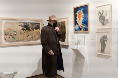 An older man wearing a coat and face mask, stands in an art fair booth in which several drawings hang on a wall.