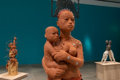 A feminine terra-cotta adult holding a clay baby. The adult has metallic circular hardware drilled into her body at various points.