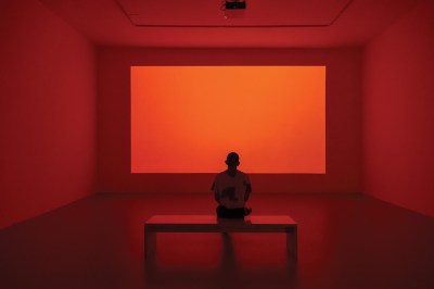A figure sitting on a bench is silhouetted by a glowing screen that fills the otherwise empty room with orange-red light.