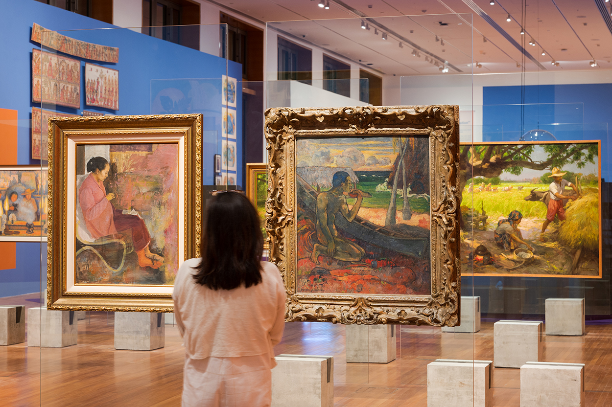 An array of colorful jewel-tone landscape paintings are displayed on vertical clear planks in a royal blue gallery.