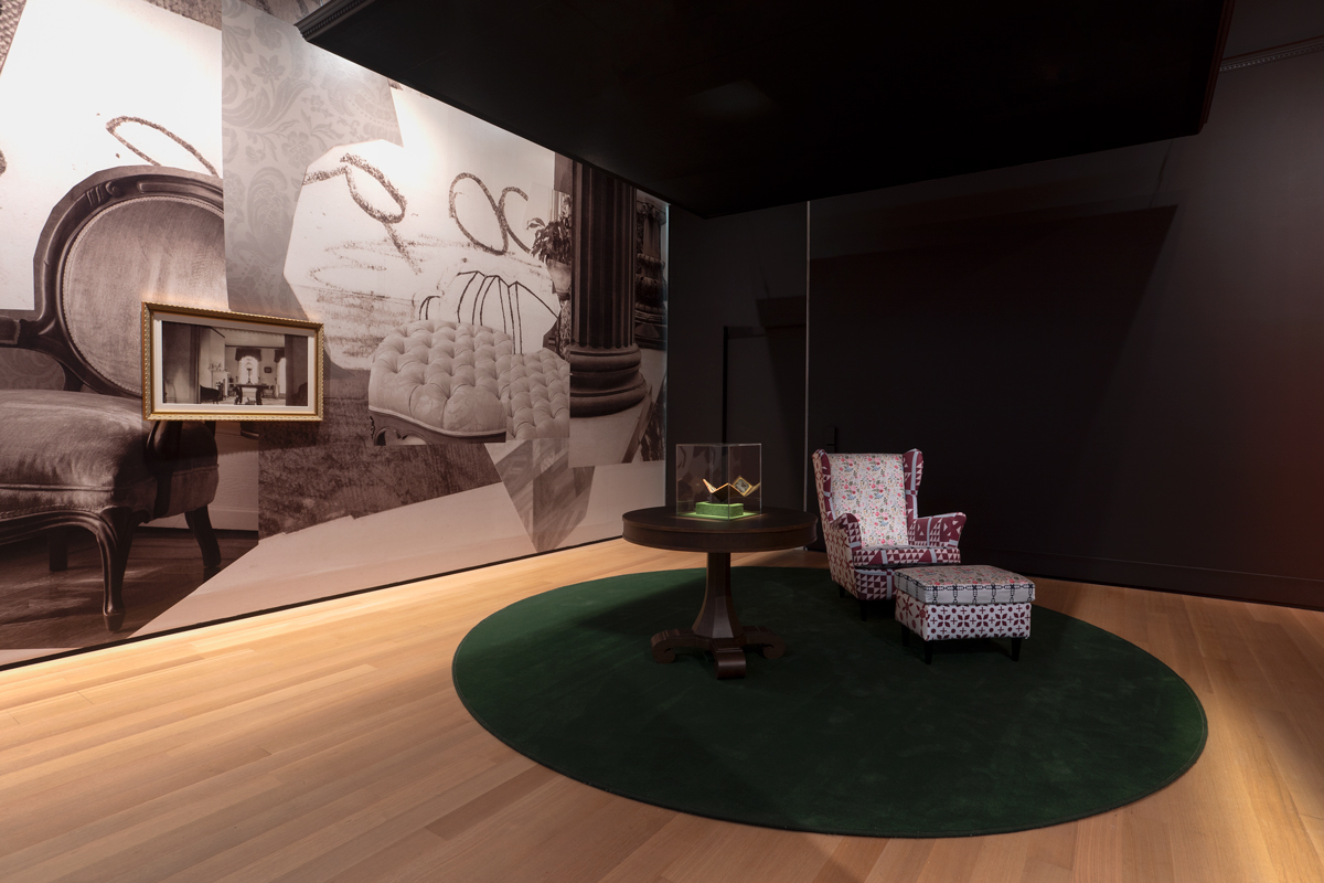 View of a museum exhibition with an arm chair and table with a miniature in a glass vitrine. Behind is a wallpaper made of various photographs of interiors.