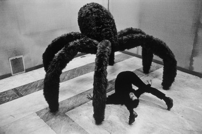 A furry sculpture of a spider looming above a man with his legs bent over his head.