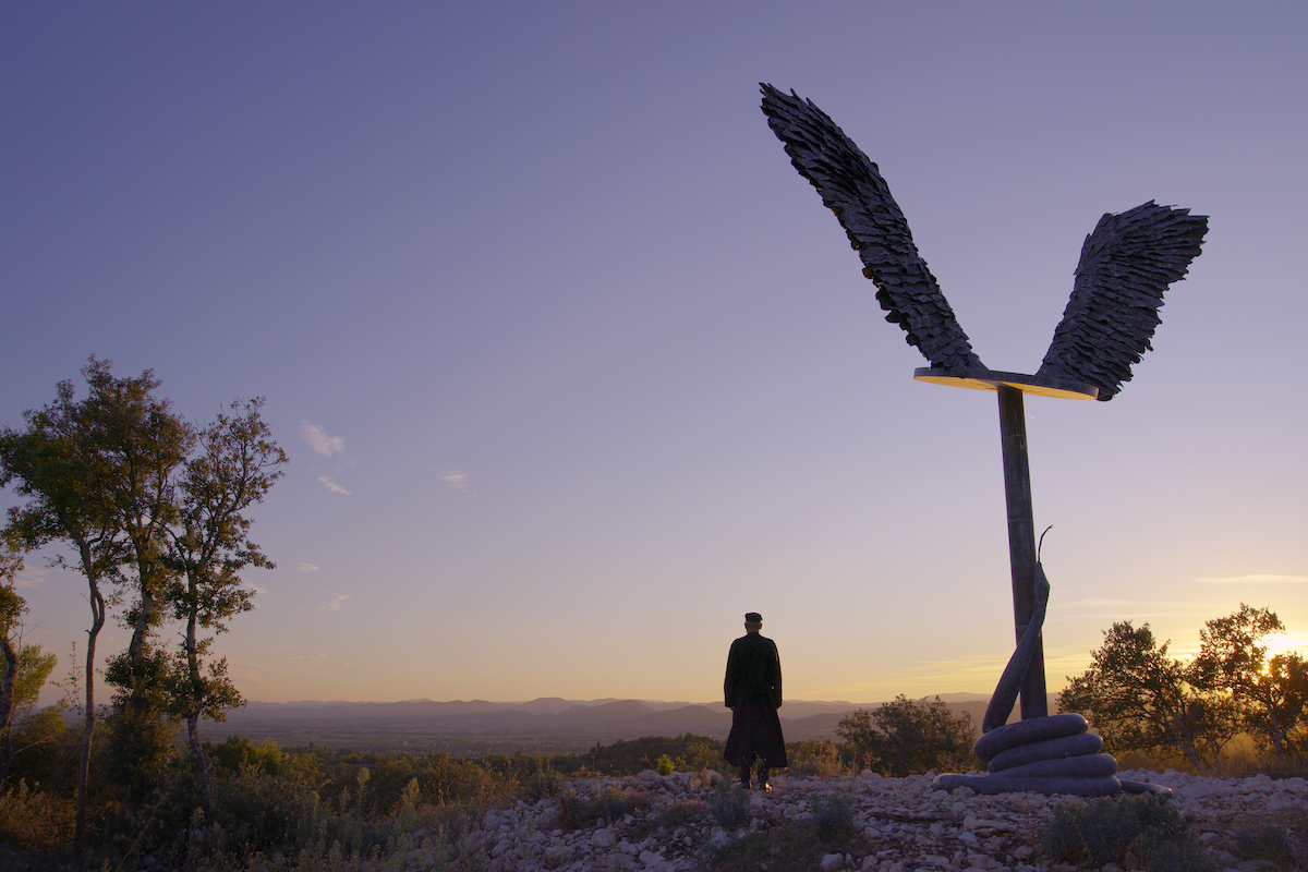 A man seen from behind standing next to a tall sculpture of a pair of angel's wings. The sun sets in the distance.