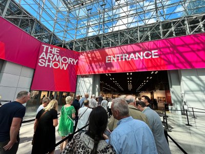 People stand in line in an airy, well-lit convention center to enter an art fair. Signage above reads The Armory Show and Entrance.