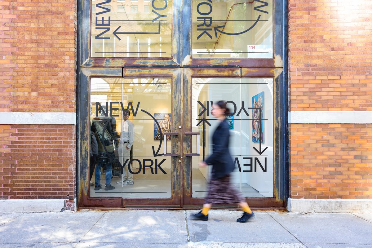 A person walks by a brick building with glass doors that have a logo that reads New York with arrows.