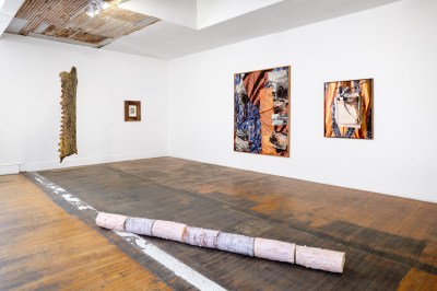 Installation view of a gallery exhibition with hardwood floors. Three works hang on the wall. One work hangs away from the walls in the left and one sculpture lays on the floor in the foregournd.