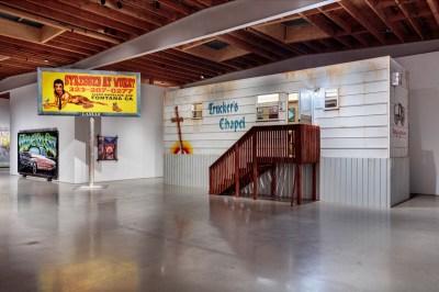 View of an art gallery with a billboard that says 'Stressed at Work?' with a nude woman and an installation that resembles a mobile home, with a wooden staircase.