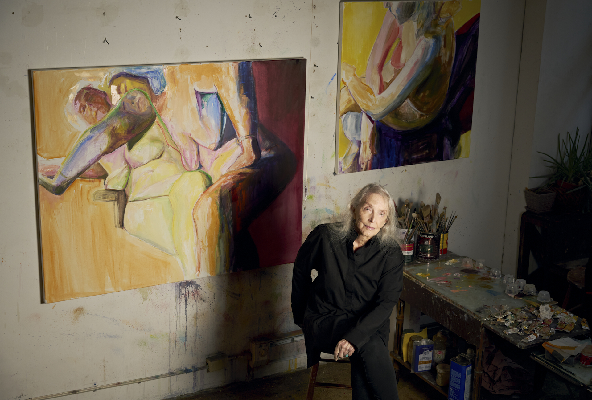 A 91 year old white woman with silver hair and blue eyes sits in front of two colorful, expressive figurative paintings. Next to her, tubes of paints and brushes fill a small table.