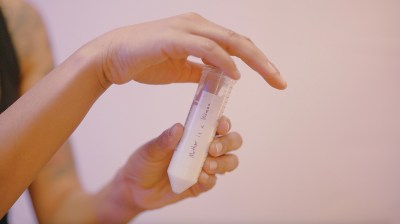 Light-skinned hands hold a test tube containing a white liquid and labeled "mother is a woman." The camera's focus is soft and the background is light pink.