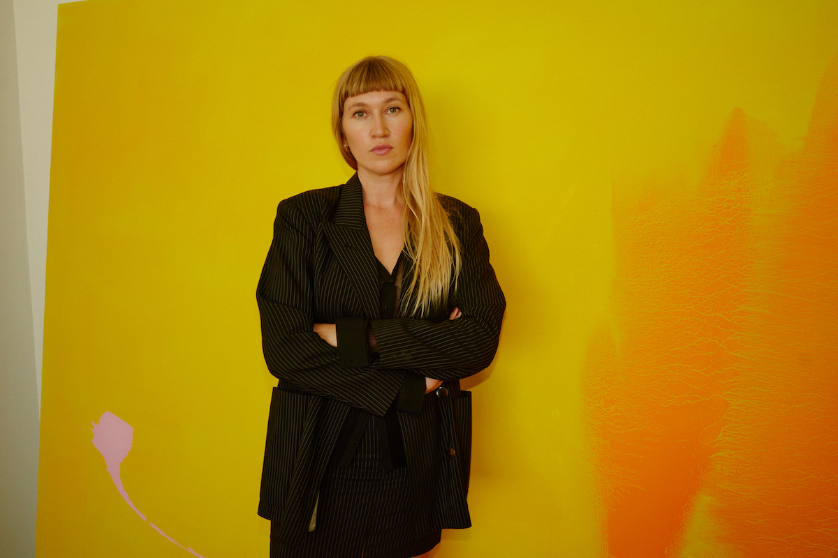 A woman with blonde bangs in a black jacket. She stands before a gigantic yellow painting holding her arms to her chest.