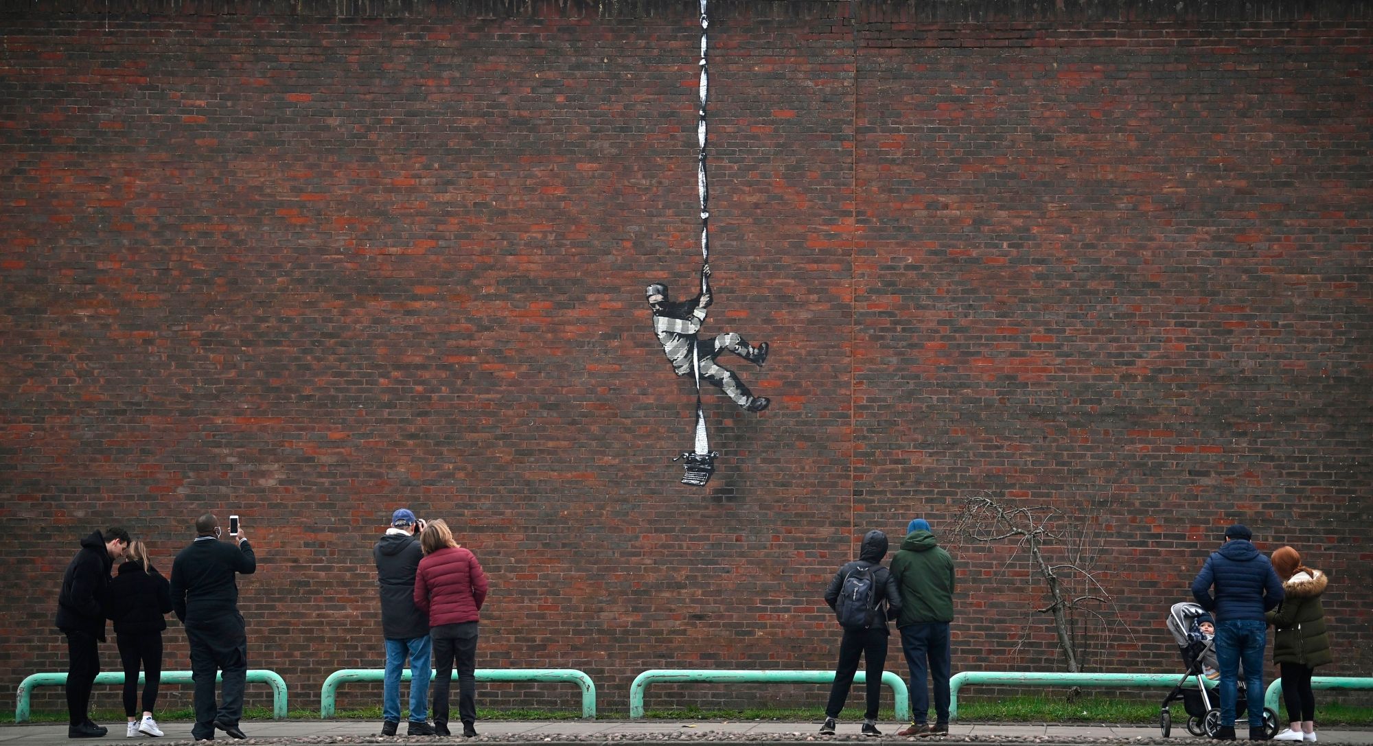 TOPSHOT - Members of the public pause to look at an artwork bearing the hallmarks of street artist Banksy on the side of Reading Prison in Reading, west of London, on March 2, 2021. - The picture shows a prisoner, possibly resembling famous inmate Oscar Wilde, escaping on a rope made of bedsheets tied to a typewriter. The work has not yet been claimed by Banksy. (Photo by BEN STANSALL / AFP) / RESTRICTED TO EDITORIAL USE - MANDATORY MENTION OF THE ARTIST UPON PUBLICATION - TO ILLUSTRATE THE EVENT AS SPECIFIED IN THE CAPTION (Photo by BEN STANSALL/AFP via Getty Images)