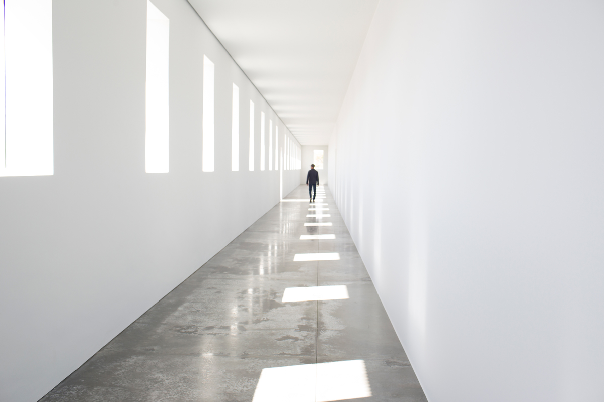 A person walks through a long white hallway that is punctuated with windows and their shadows.