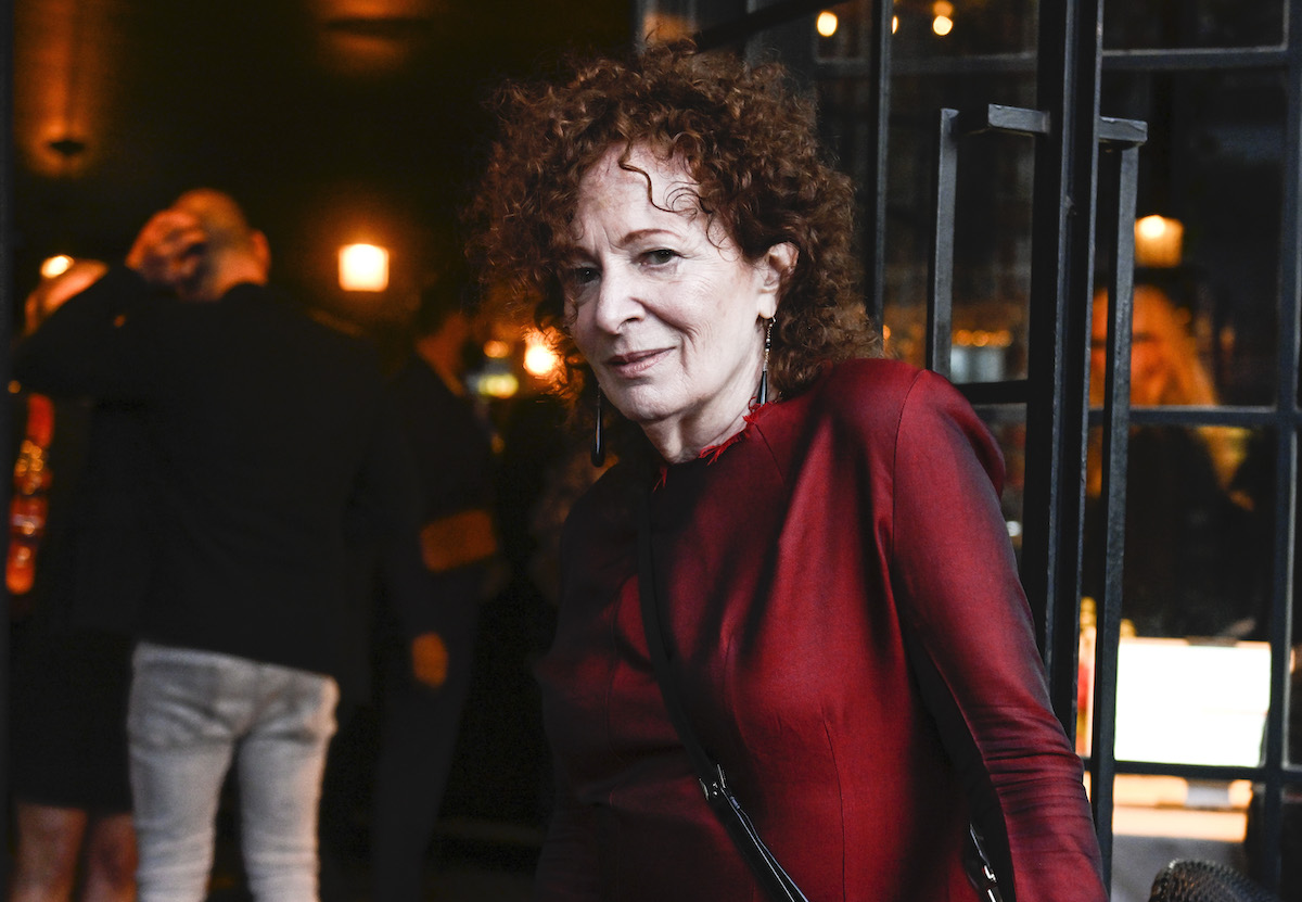 A woman with curly red hair wearing a red top and a purse. She stands before others at a party.
