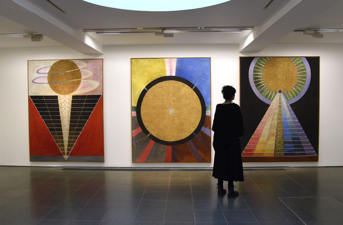 A woman staring at three large paintings with pyramidal forms topped with orbs.