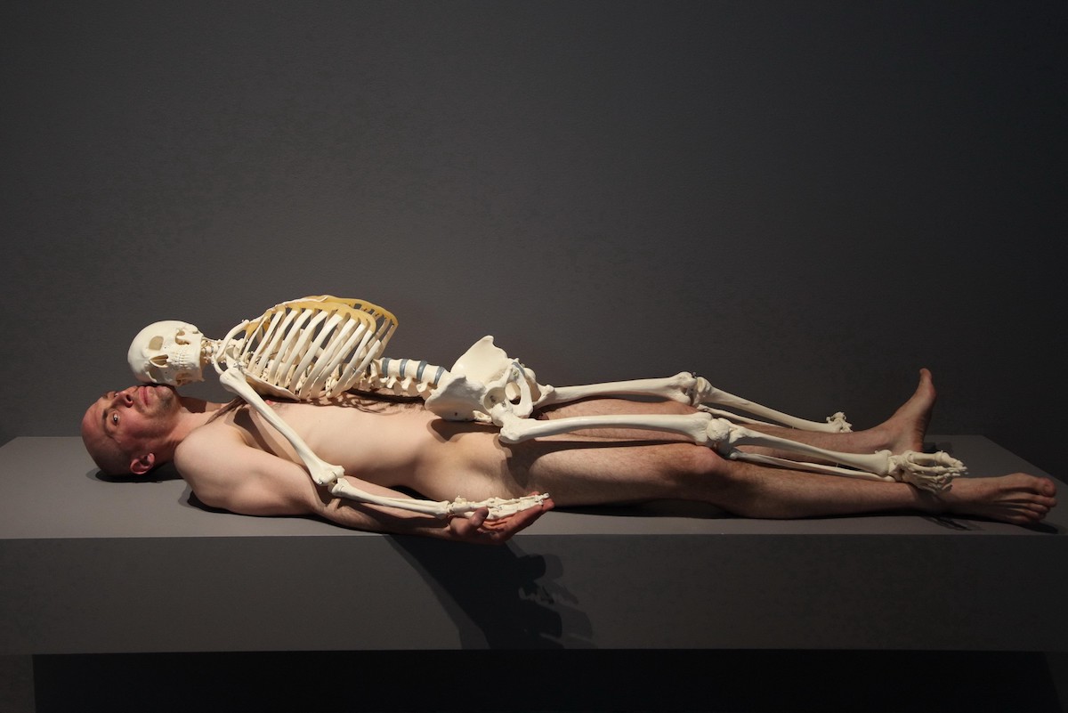 A nude white man lying beneath a skeleton.