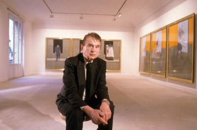 A white man seated in a gallery with paintings around him.