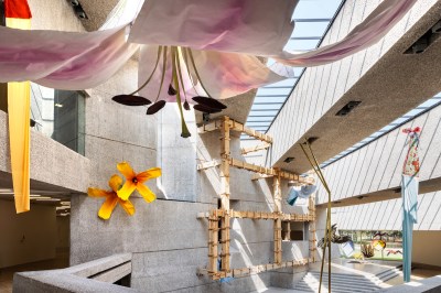 A giant sculpture of a flower hanging from the ceiling of an open atrium whose walls are also hung with large flowers and a structure recalling the wooden armature of a house.