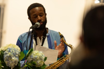 A black man sits at a microphone; hydrengas partially eclipse the view. His eyes are closed as if taking a poignant pause as a saxaphone sits in his lap.