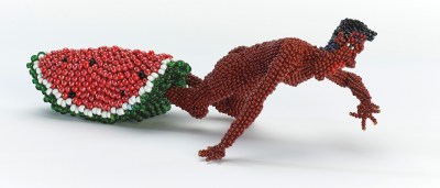 A beaded sculpture showing a nude Black man crawling a way from a giant slice of watermelon that engulfs his feet.