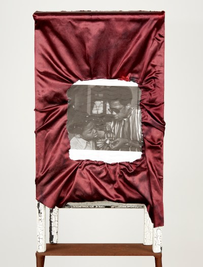 A photograph of a Black girl and a Black man attached to a piece of crimson fabric that covers a white table whose legs are cracked.