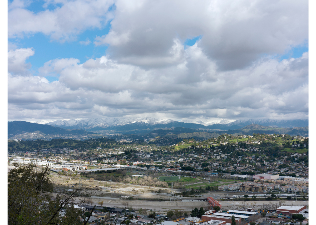 A photograph showing a view of Los Angeles from faraway with a cloudy sky and snow-capped mountains.