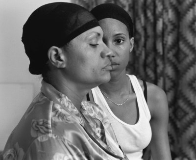 Two Black women posed so that one is shown from the side and the other is shown dead-on, staring at the camera. The one facing away has her eye closed and wears a floral-print shirt. They are photographed before a curtain.