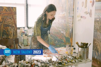 A young woman in a patterned shirt and paint-spattered apron wearing blue gloves holds a palette knife and leans over a table covered with tubes of paint