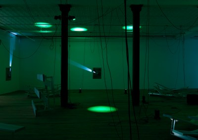 An installation view shows a space with dim green lighting in which several spotlights are pointed at the floor, walls, and ceiling.