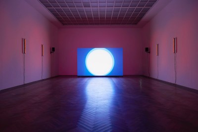 A vast gallery with purple lighting. At its center, on the floor, is a screen showing what looks like a close-up of the sun against a blue sky.