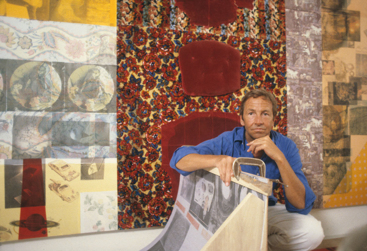 Portrait of Robert Rauschenberg posing in front of a large-scale artwork that is made up of assemblaged elements