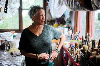 a woman from the waist up, looking off to the right, in a dark v-neck shirt, standing in the foreground of an art studio with various art-making materials filling the space