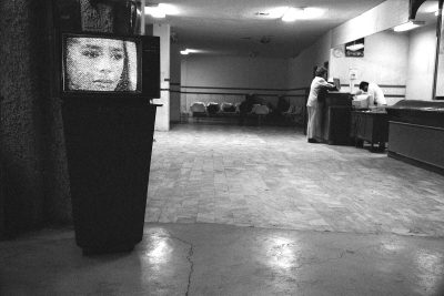 A black-and-white photograph of a waiting room with a person at a desk. A TV set closer to the camera plays a pixelated image of a woman's face.
