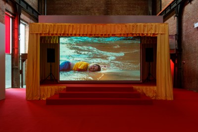 In a red-carpeted room, a small stage contains a video projection depicting a young child lying down on the sand at a shoreline.