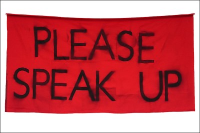 A red piece of fabric has smudgy text in all caps that reads: please speak up.