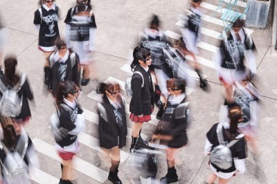 An East Asian woman with a long ponytail is wearing sunglasses, a blazer, and a plaid skirt. She's standing on a cross walk and women in similar outfits surround her, blurred out as if moving quickly.