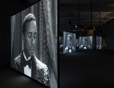 A darkened room with five large video screens arranged at various angles. Each one features the same image of a black man from the chest up, wearing a tuxedo, his eyes closed and falling snow collecting on his hair and shoulders.