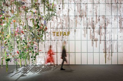 Two blurred figures walk past a wall that says TEFAF. At left is a suspended installation containing various flowers and other plants.