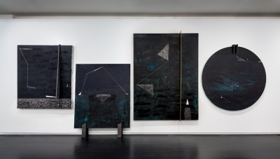 Four textured black paintings hang in a white cube. 3 are rectangular; one is resting on the ground. The 4th is circular.