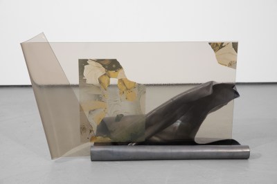 A glass sheet stands upright, with its left corner folded over; it is supported by a rolled metal sheet. An illegible image comprising mostly amberish hues has been transferred on to the glass, roughly in the center, with one corner ripped off and displaced to the right side.