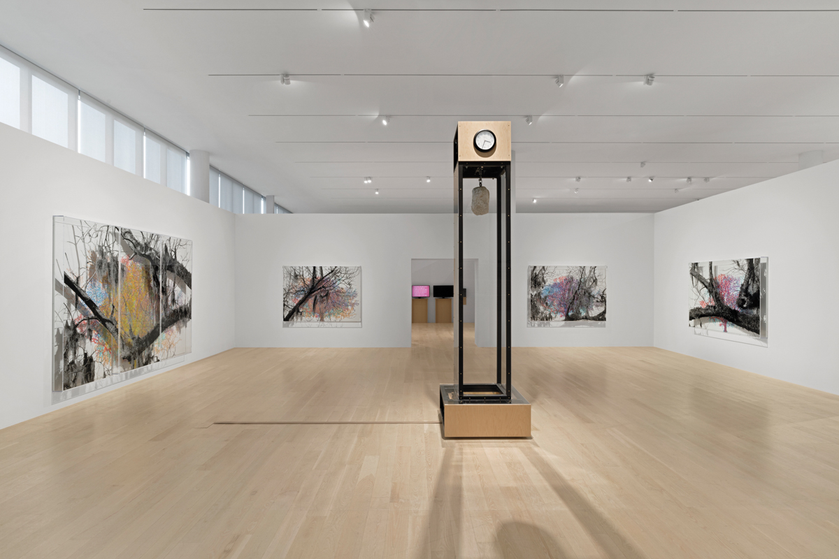 Installation view of a museum exhibition with a large sculpture that looks like a grandfather clock with a piece of granite suspended in its center. On the walls are artworks showing black-and-white details of trees over grids.